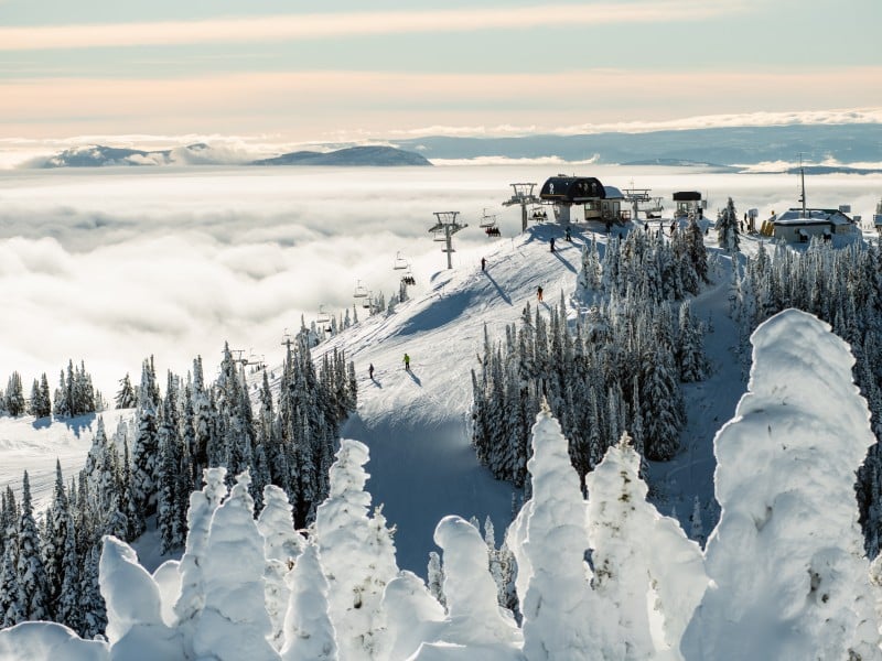 Thick fog rolling in below the crest of a snowy mountain as a chairlift operates.
