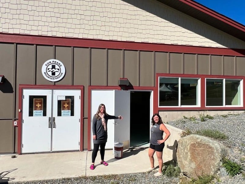Two women present the far right of a health centre with white doors and brown siding.