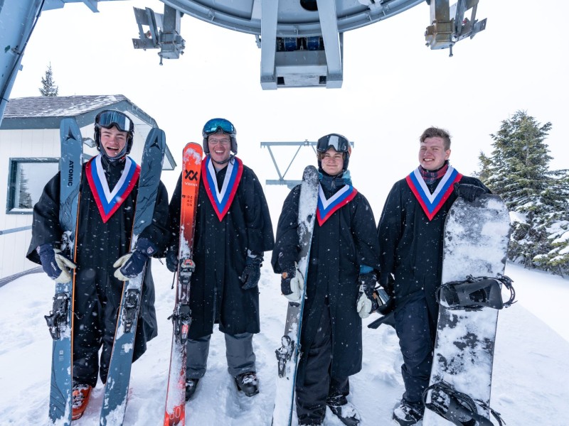 2022 graduates of Sun Peaks Secondary Academy pose with a wintry background holding skis and wearing graduate robes.