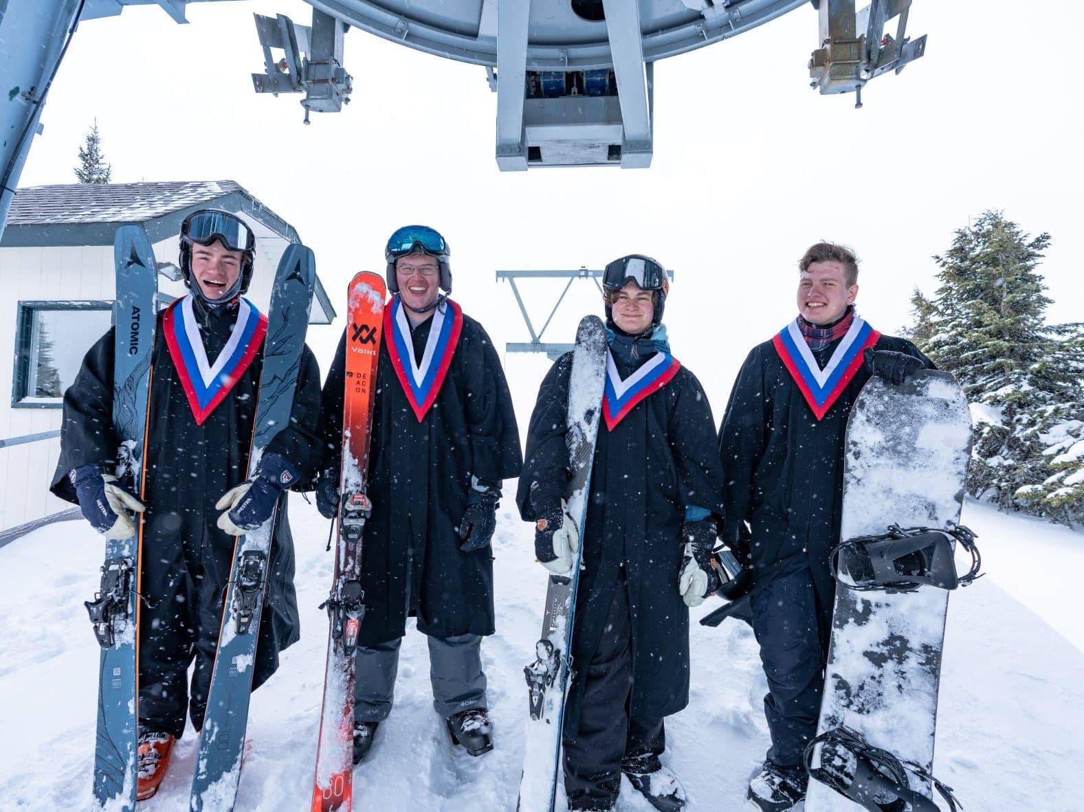 2022 graduates of Sun Peaks Secondary Academy pose with a wintry background holding skis and wearing graduate robes.