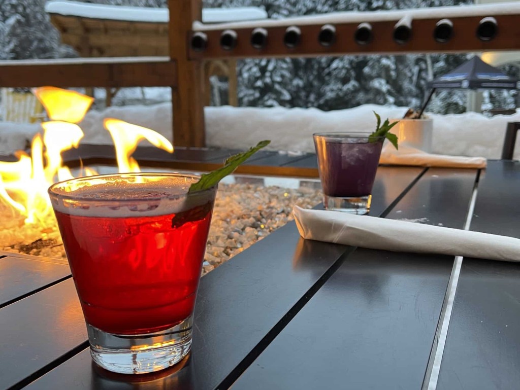 On the patio beside a tabletop fire, two cold drinks and one warm drink stand in front of a snowy backdrop.