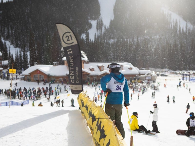 Overlooking a crowd at the village base, the shot focuses on a ski racer by a gate and sign.