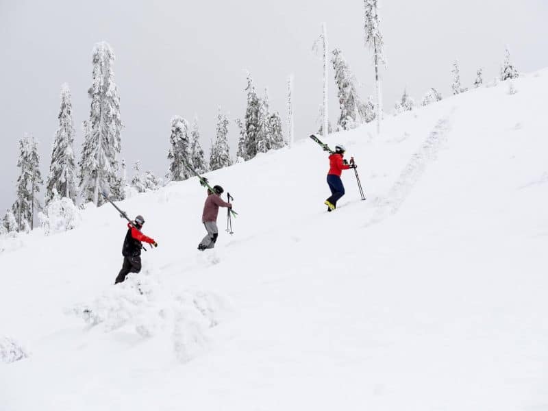 Three skiers hike up a mountain carrying their skis, side view.