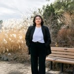 Girl with dark hair stands in front of a bench with a black blazer and white tee shirt. She is wearing black pants and there are plants in the back ground. It is sunny and cloudy.