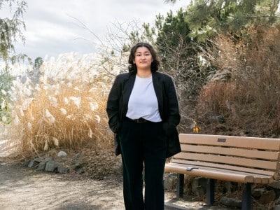 Girl with dark hair stands in front of a bench with a black blazer and white tee shirt. She is wearing black pants and there are plants in the back ground. It is sunny and cloudy.