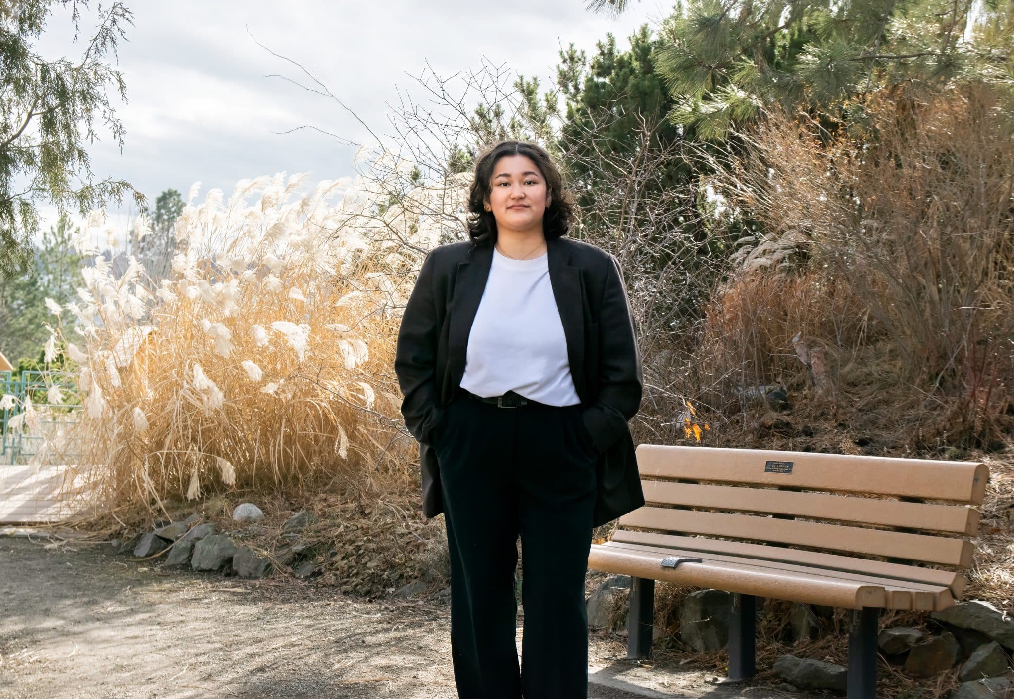 Girl with dark hair stands in front of a bench with a black blazer and white tee shirt. She is wearing black pants and there are plants in the back ground. It is sunny and cloudy.